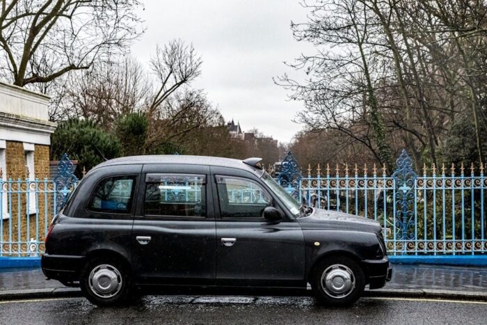 Black London-style taxi parked beside an ornate blue fence on a wet street